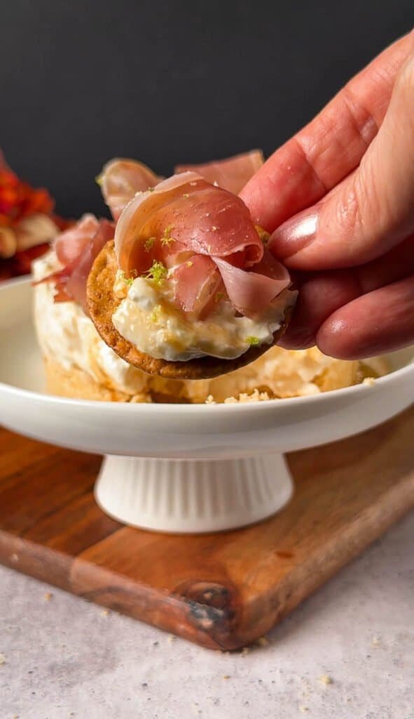 Hand holding a cracker topped with pear cheesecake filling and prosciutto