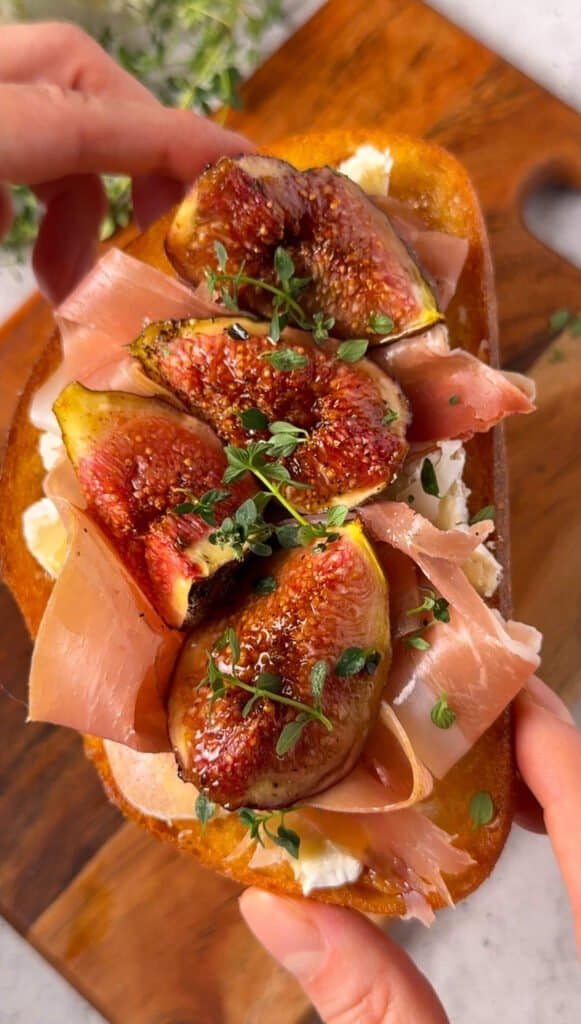 Hand holding a roasted fig and brie toast topped with prosciutto and fresh thyme