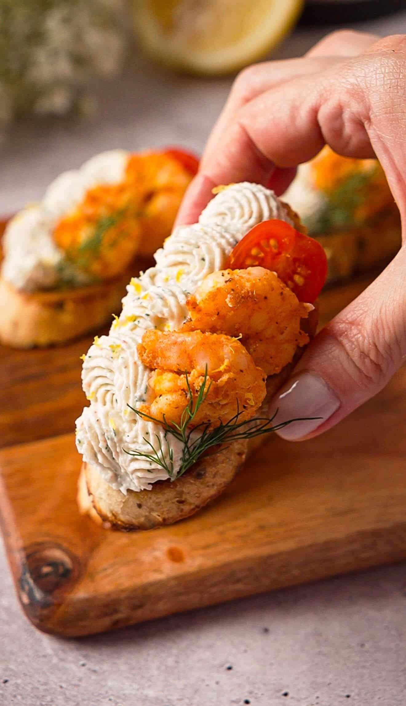 Close-up of a hand holding whipped crab crostini topped with garlic shrimp and cherry tomato