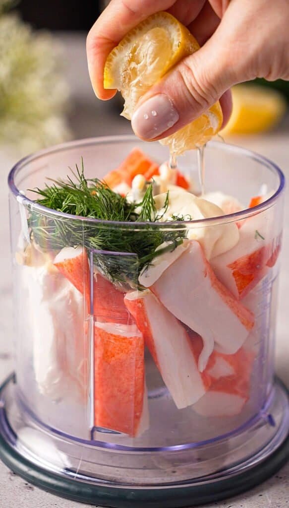 Lemon being squeezed into a food processor with imitation crab, mayonnaise, and fresh dill