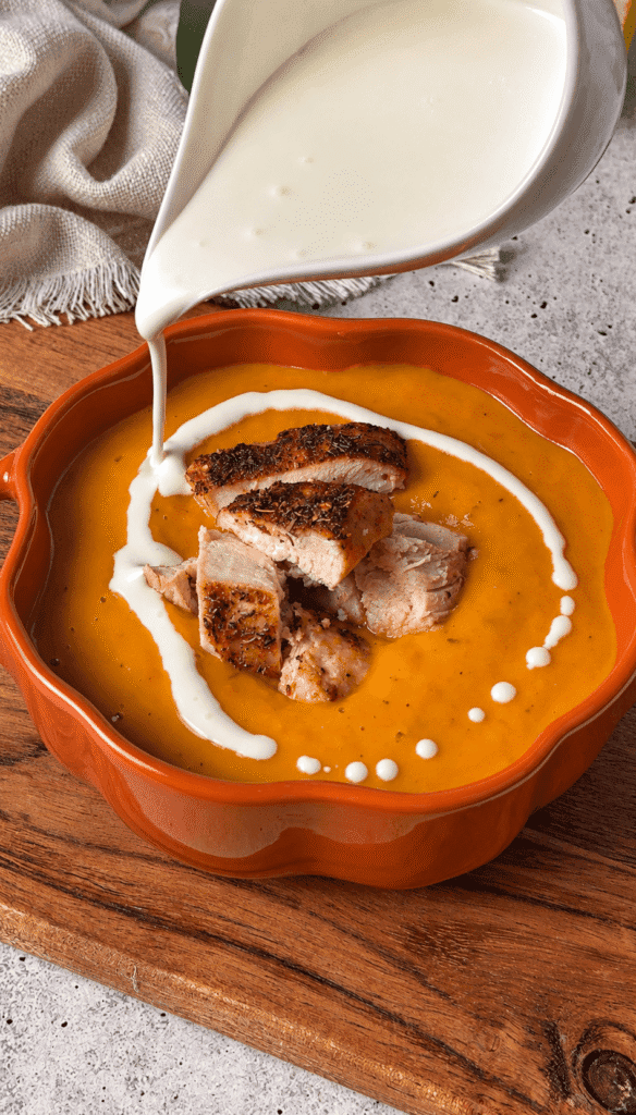 Heavy cream being drizzled over butternut squash soup topped with roasted chicken