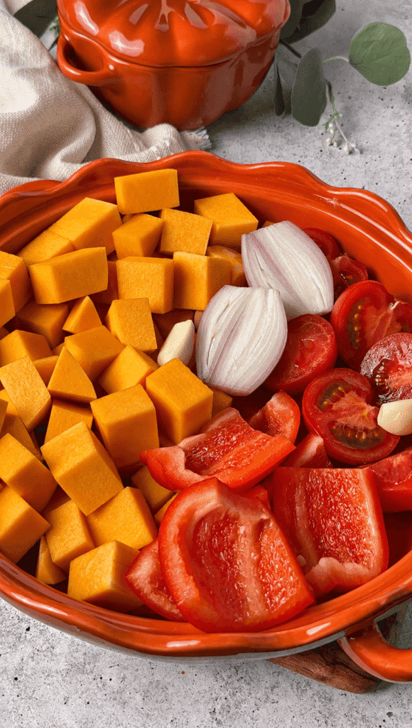 Cubed butternut squash, red bell pepper, tomatoes, shallot and garlic in an orange baking dish
