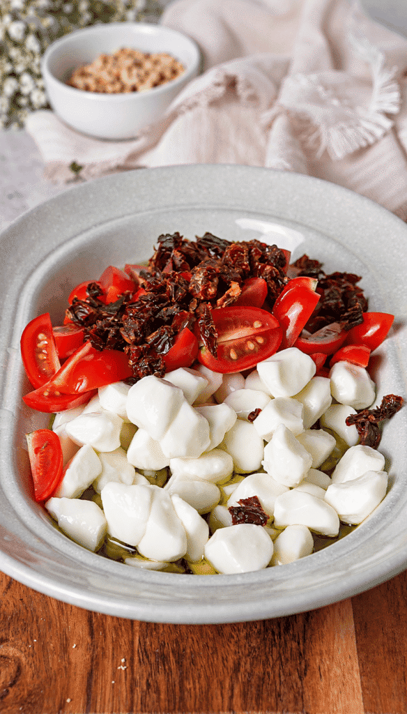 Mozzarella pearls, cherry tomatoes, and chopped sun-dried tomatoes arranged in a bowl with olive oil.