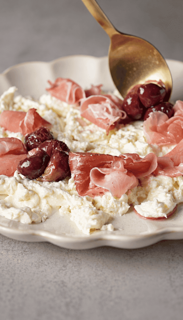 Spooning roasted red grapes over creamy burrata and folded prosciutto on a scalloped platter