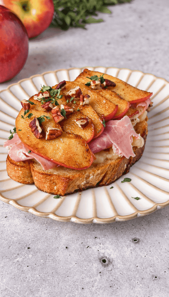 Apple Brie toast with ham and overlapping caramelized apple slices, finished with pecans and thyme on a white plate.