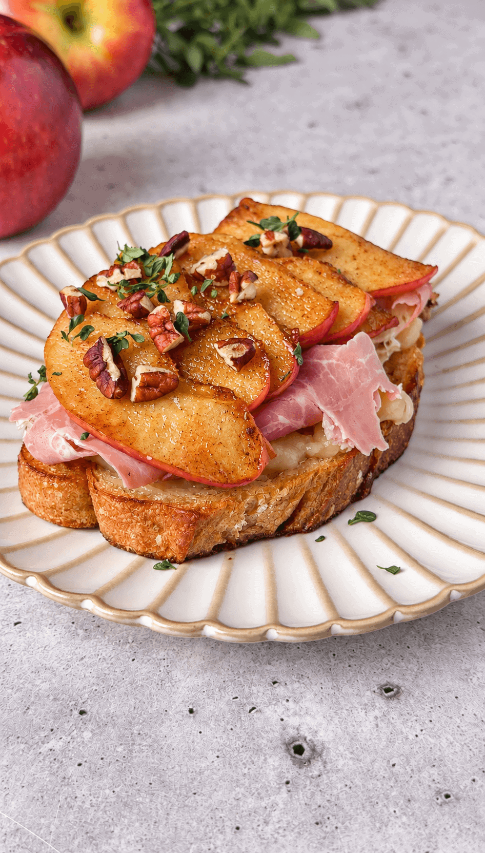 Apple Brie toast with ham and overlapping caramelized apple slices, finished with pecans and thyme on a white plate.