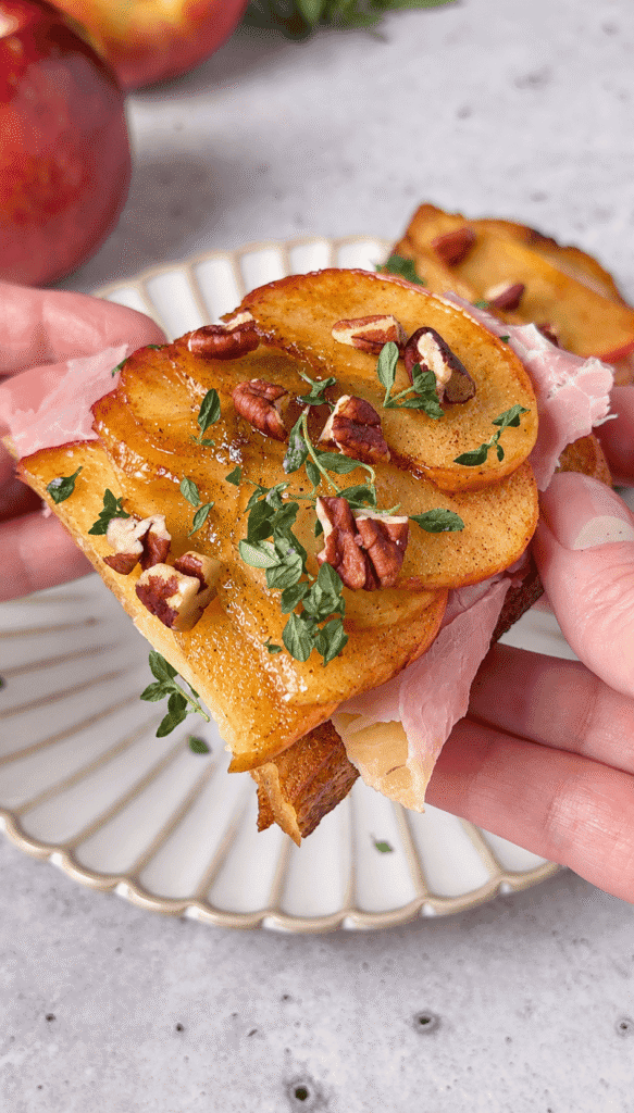 Hands holding apple Brie toast topped with ham, caramelized apples, chopped pecans, and fresh thyme.