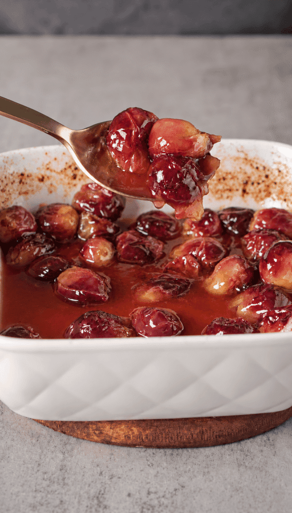 A gold spoon lifting softened balsamic-roasted red grapes above a white baking dish filled with their juices.