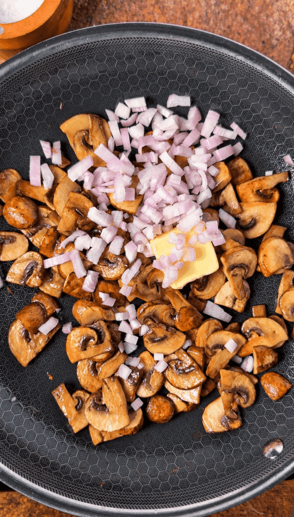 Browned cremini mushrooms in a skillet with chopped shallots and a pat of butter.