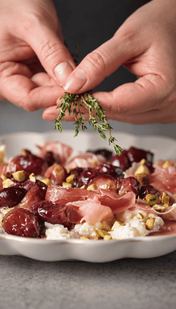 Hands stripping fresh thyme over burrata topped with roasted grapes, prosciutto, and chopped pistachios.