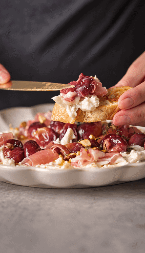Toasted bread topped with creamy burrata, roasted grapes, and prosciutto, held above the serving platter.