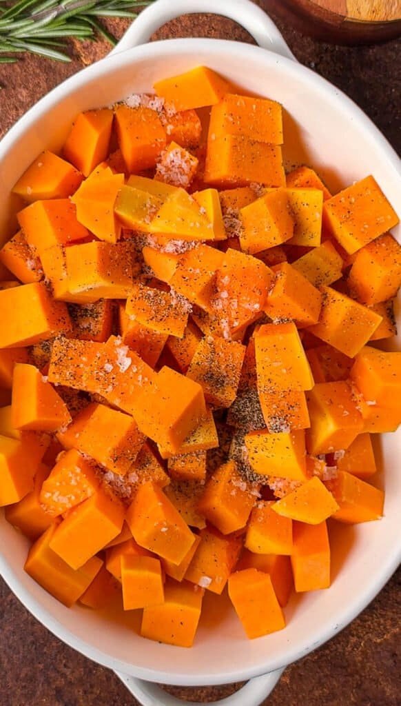 Raw butternut squash cubes seasoned with salt and black pepper in a white baking dish