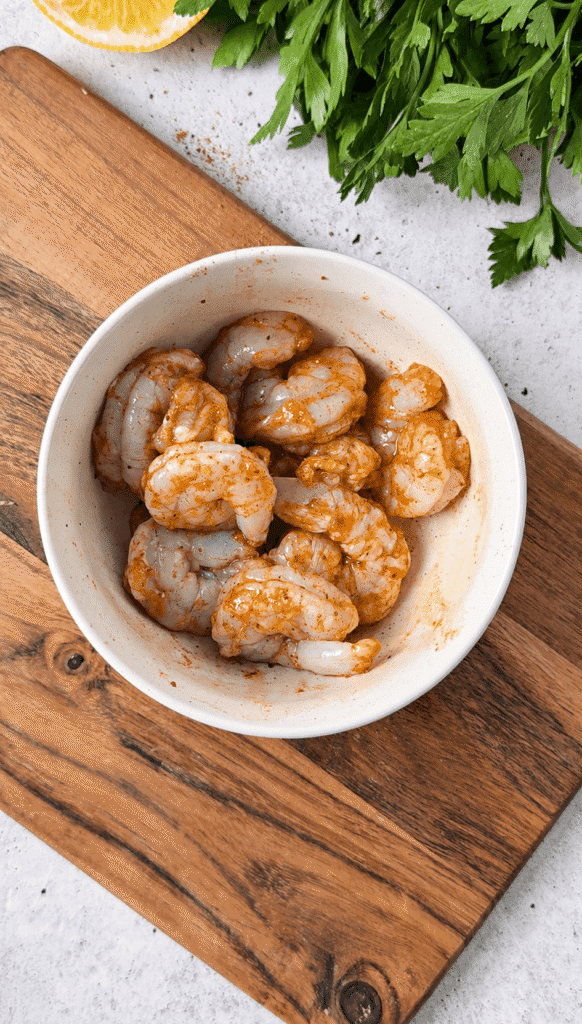 Raw peeled shrimp coated in Cajun seasoning in a white bowl on a wooden board.