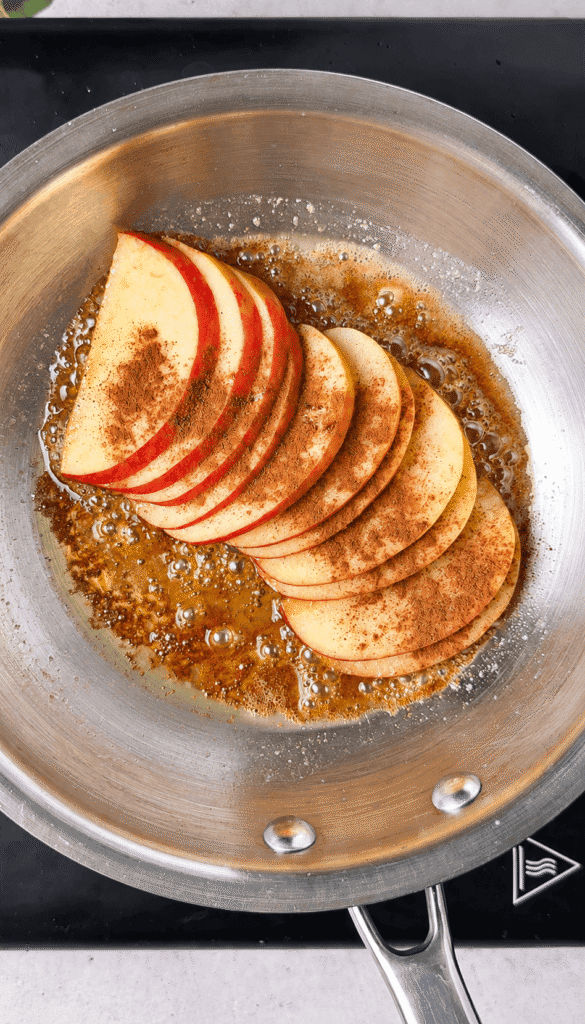 Red-skinned apple slices sprinkled with cinnamon in bubbling butter and brown sugar in a stainless steel skillet.