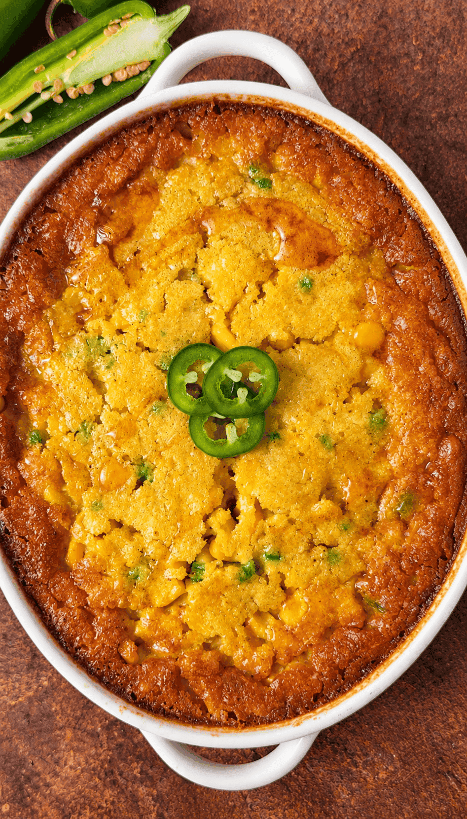 Golden baked corn pudding with a honey drizzle and fresh jalapeño slices in a white oval dish.