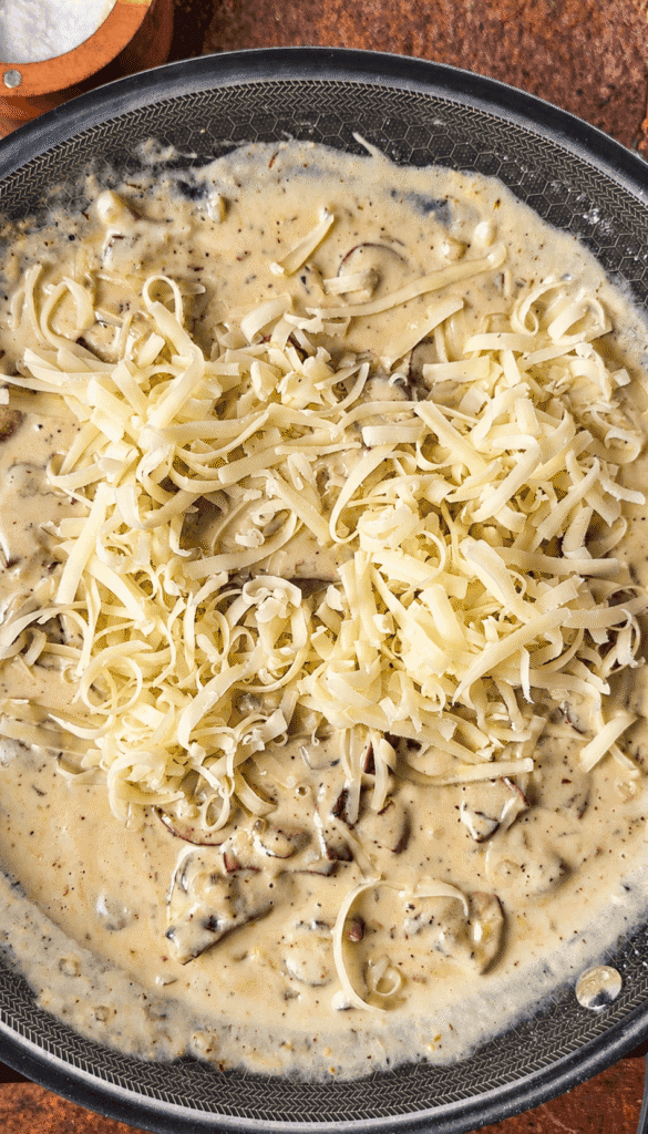 Grated Gruyère piled over creamy mushroom sauce in a skillet, ready to stir in.