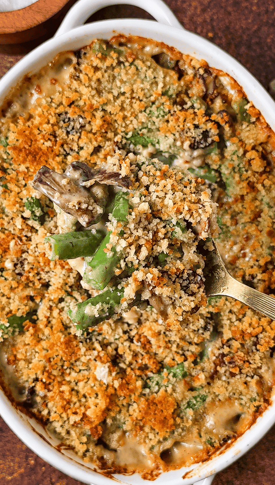 A gold spoon lifts green bean casserole with mushrooms, creamy Gruyère sauce, and golden breadcrumbs from a white baking dish.