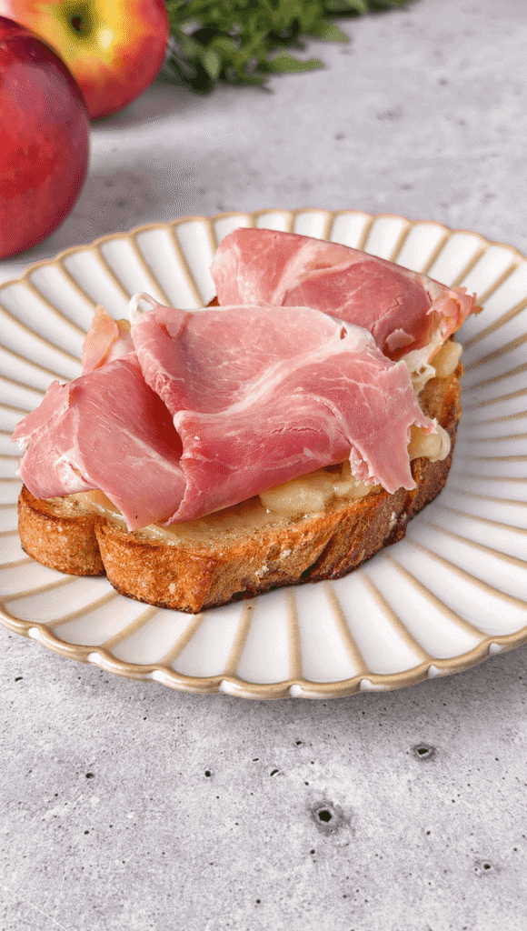 Toasted sourdough layered with melted Brie and thinly folded ham on a white scalloped plate.