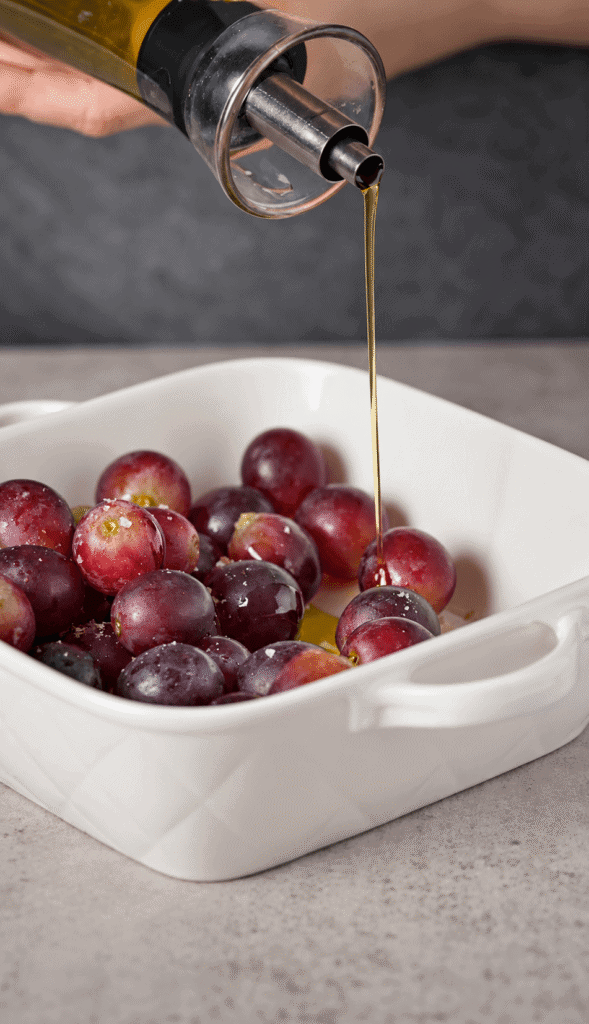 Olive oil being drizzled over seasoned red grapes in a white baking dish before roasting.