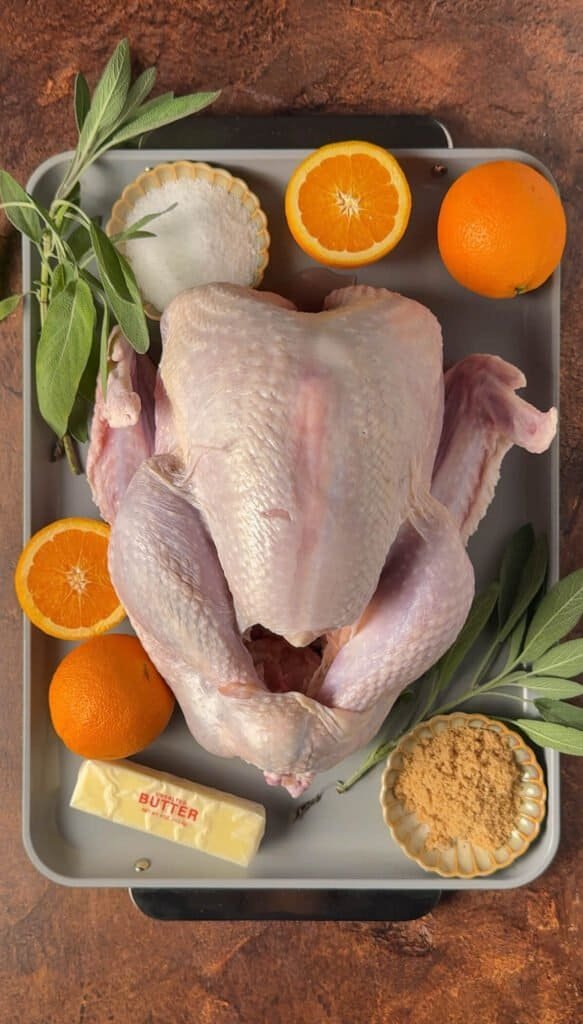 Raw whole turkey in a roasting pan surrounded by fresh oranges, sage, kosher salt, brown sugar, and butter.