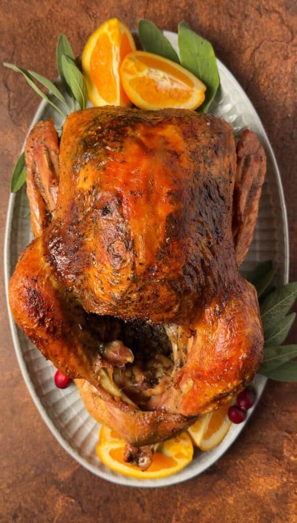 Whole roasted citrus turkey with golden brown skin, garnished with fresh orange wedges, sage leaves, and cranberries on a serving platter.
